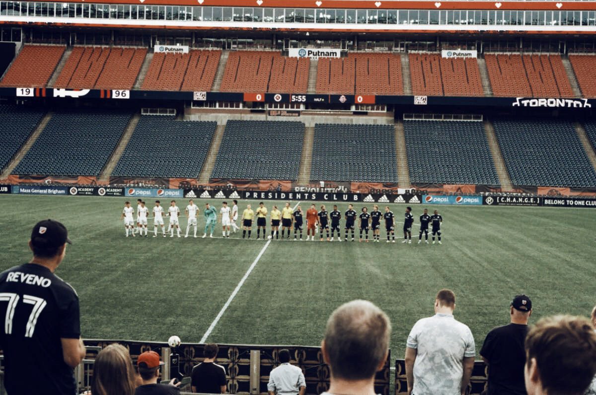 Two soccer teams lined up on the pitch before kickoff, in a stadium with empty stands